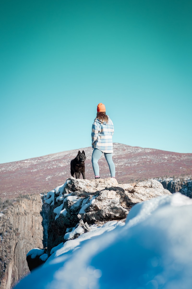 woman in white jacket and white pants standing on rocky hill with black dog during daytime